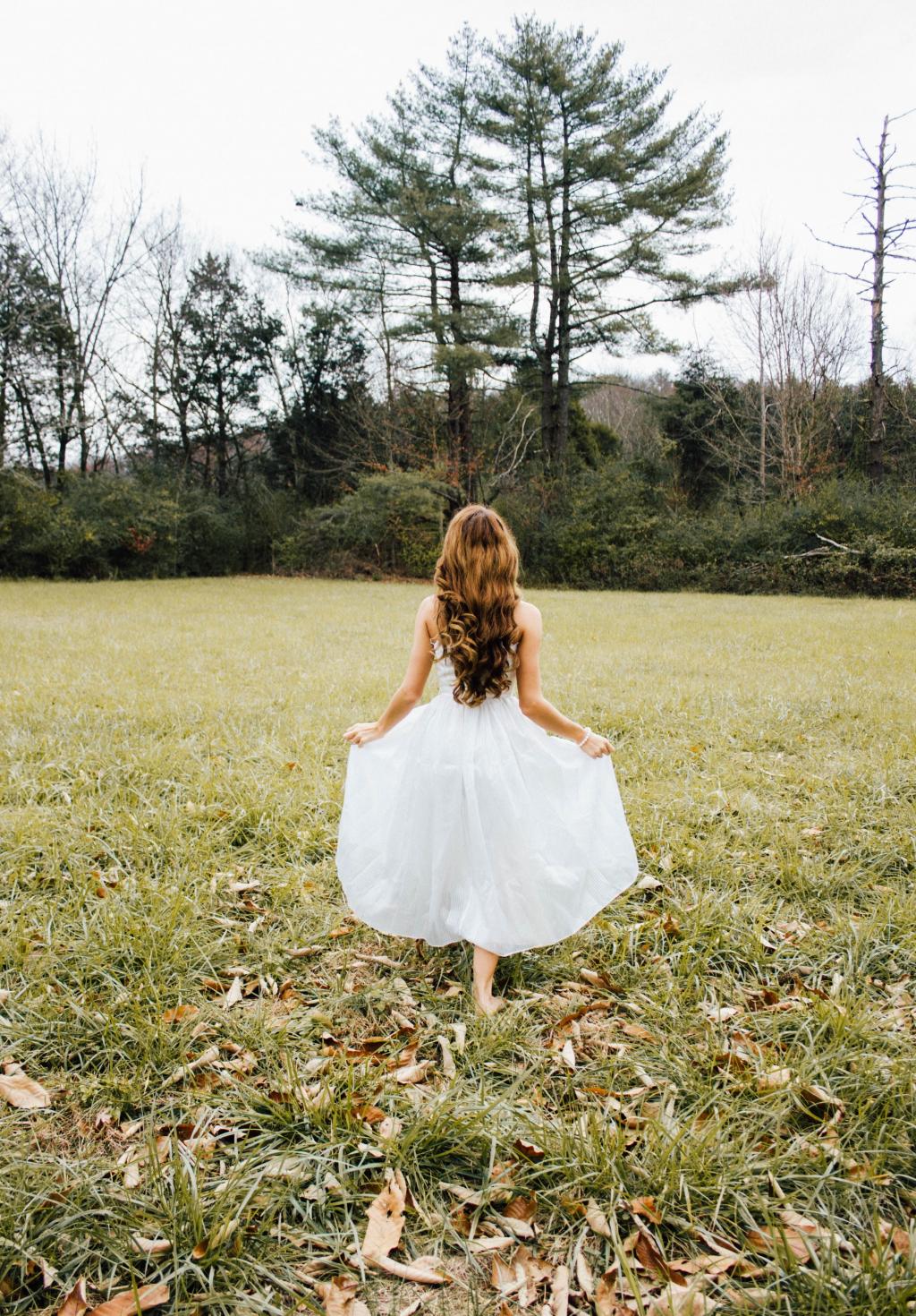 站在草地上的白色连衣裙的女孩,同时面对高大的树木,在一天时间高清壁纸