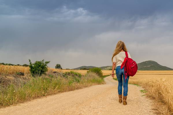 女人穿着白色上衣和蓝色粗斜纹棉布牛仔裤配红色背包走在棕色的草地上,在白天高清壁纸附近的岩石道路上