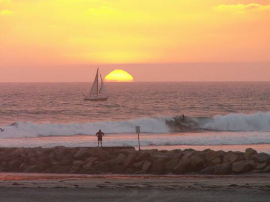人站在海洋与帆船在日落期间的岩石上高清壁纸